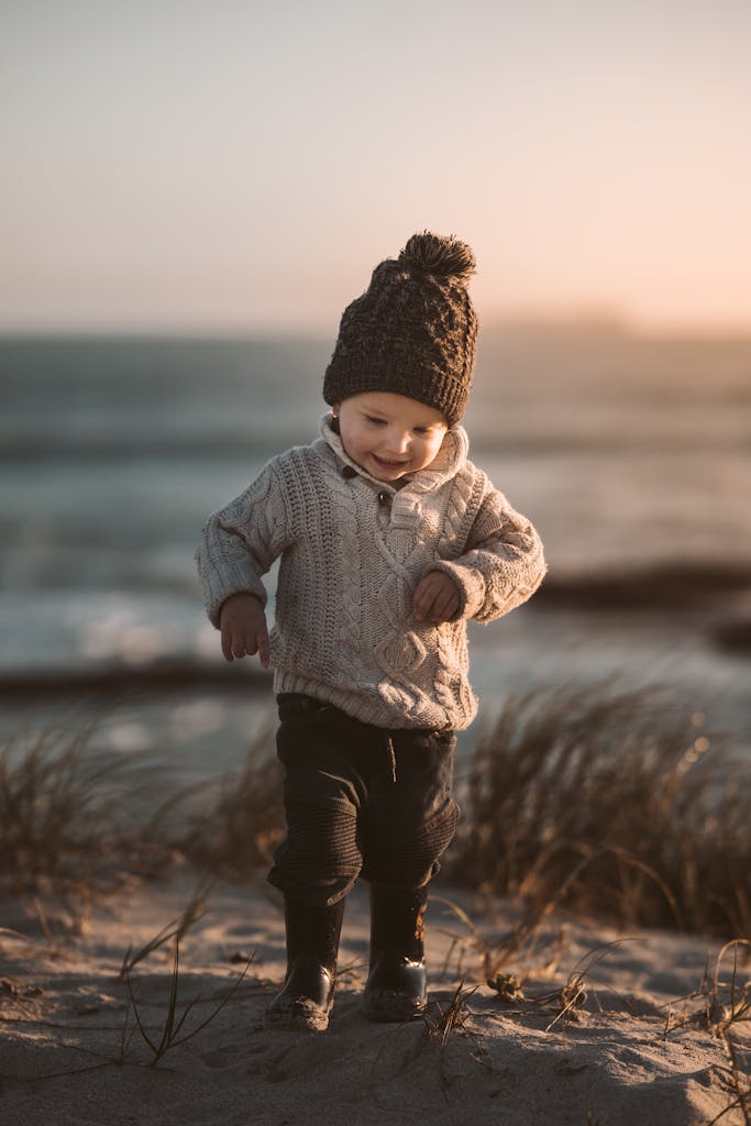 Adorable toddler in warm knitwear exploring a sandy beach with a joyful smile at sunset.