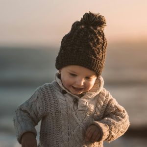 A smiling toddler in a knitted sweater and winter hat enjoying a day by the sea at sunset.