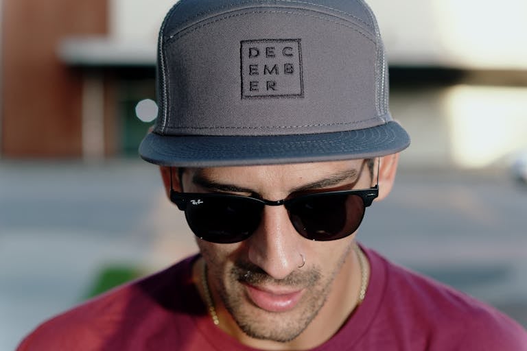 Close-up portrait of a man wearing a cap labeled 'December' and sunglasses. Outdoors and stylish.