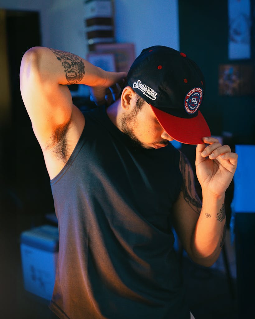 Man in casual wear adjusting cap, showcasing tattoos in moody lighting indoors.